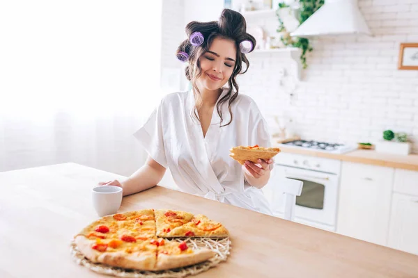 Jovem feliz olhar para pizza fatia na mão e sorrir. Use encrespadores no cabelo e vestido de vestir branco no corpo. Sozinho na cozinha. Alimentos gordurosos . — Fotografia de Stock