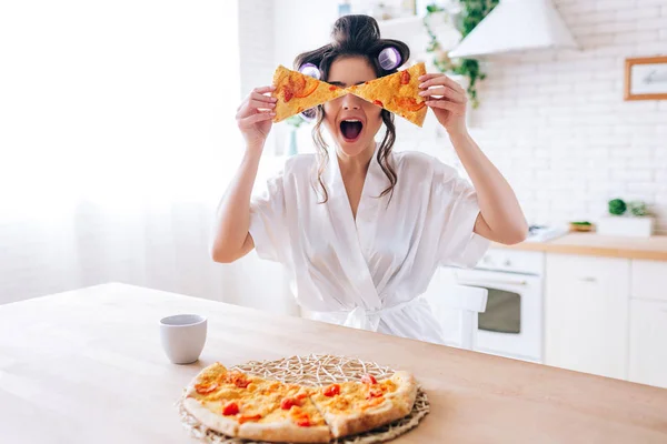 Jovem feliz entusiasmada na cozinha entranhando com fatias de pizza. Cobrir os olhos com comida. Empregada doméstica despreocupada muito brincalhão. Usa um roupão branco. Descuidada dona de casa . — Fotografia de Stock