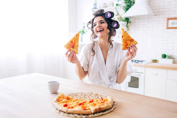 Positive emotionale junge unbeschwerte Haushälterin in der Küche. Zwei Scheiben Pizza in der Hand und lächeln. Tasse auf dem Tisch. Allein in der Küche. Leben ohne Arbeit. — Stockfoto