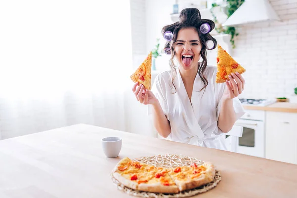 Lekfull ung kvinna visar tungan på kameran. Hushållerska håller två skivor pizza i händerna. Kopp på bordet. Ljust dagsljus på morgonen. Livsstil utan arbete. — Stockfoto