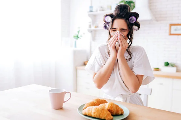 Sjuk ung kvinna täcka näsa med vävnad. Influensa. Croissant och kopp drink på bordet. Stå i köket. Ensam hem. Hemmafru leva slarvig liv. — Stockfoto