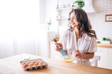 Young woman with dark hair stand in kitchen and cooking. Blending eggs. Alone. Morning daylight. Looking straight and smile. Phone on table.