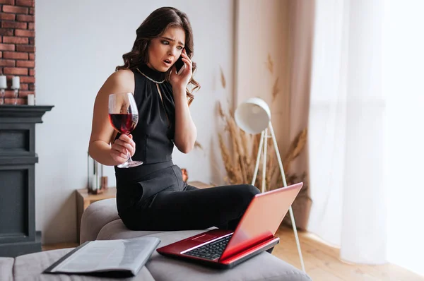 Young businesswoman work at home. Sitting on table and talking on phone. Hold glass of red wine in hand. Brunette model wear black dress and brown shawl.
