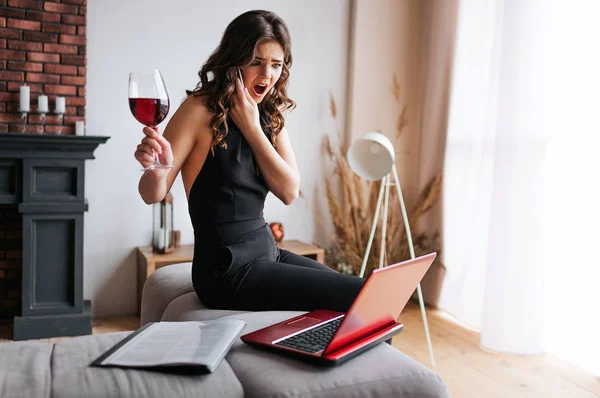 Young businesswoman work at home. Talking on phone and sitting on table in living room. Look on lpatop and hold glass woth red wine in hand. Beautiful brunette working hard.