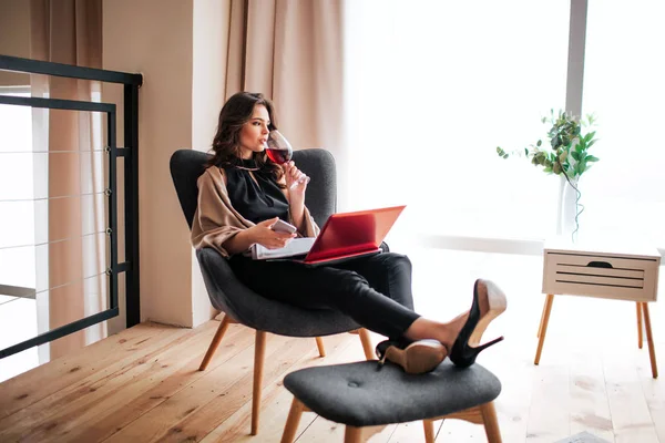 Young businesswoman work at home. Drinking red wine from glass and look to right. Hold phone in hands. Journal and laptop on legs. Remote work. Alone in room. Relaxing.