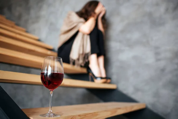 Ung kvinna har problem med alkohol. Sitter på trappan och gömmer ansiktet. Fashionabla kvinna lider av baksmälla. Ensam i fara. Berusad. Glas rött vin i Fron av bild. — Stockfoto