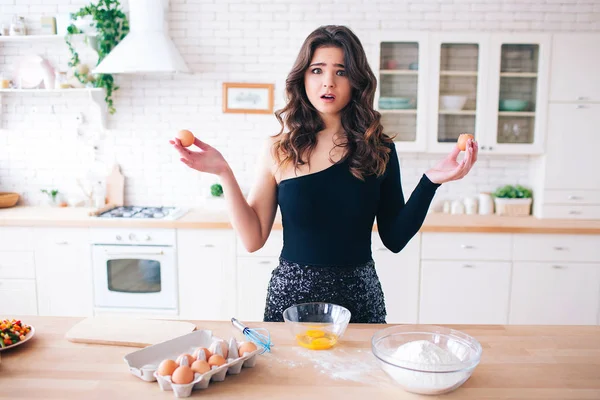 Jovem mulher bonita cozinhar comida. Ovos e farinha em talbe. Modelo confuso emocional distraído olhando diretamente para a câmera. Stand na cozinha . — Fotografia de Stock