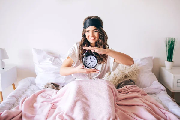 Morena joven y hermosa de pelo oscuro se despierta en su cama. Alegre mujer agradable sosteniendo reloj en las manos y sonrisa. Mira directamente a la cámara. Máscara de dormir en la frente. Modelo feliz positivo en el dormitorio . —  Fotos de Stock