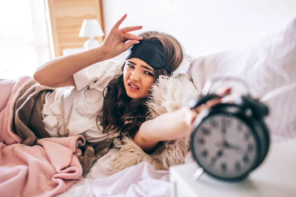 Morena joven y hermosa de pelo oscuro se despierta en su cama. Mujer irritada mintiendo y acercándose al reloj. Sostén la mano sobre la máscara para dormir. Solo en el dormitorio . —  Fotos de Stock