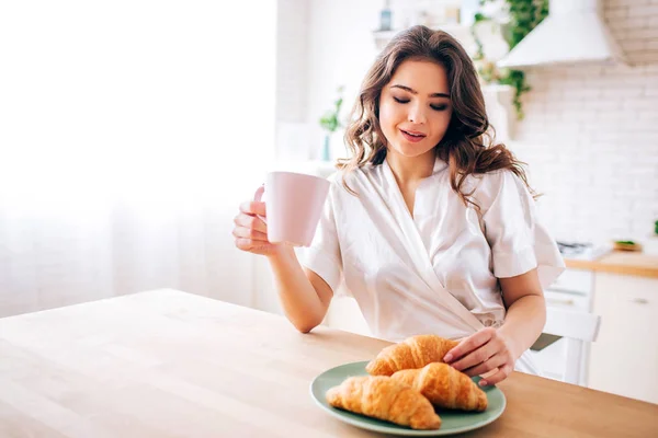 Young woman with dark hair Sitting in kitchen and drinking coffee in morning. Taking one croissant and smile. Delightful model. — Stockfoto