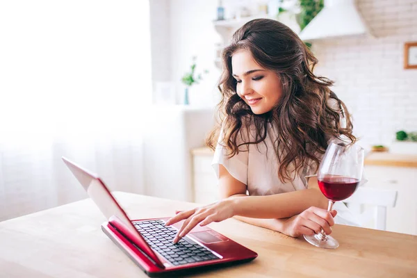 Femeie tânără cu păr negru în picioare în bucătărie și de lucru. Tastarea pe tastatura laptopului. Sticlă cu vin roşu pe masă. Singur în bucătărie . — Fotografie, imagine de stoc