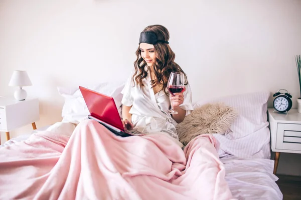 Young woman with dark hair sitting on bed in bedroom and watching movie on laptop. Alone. Drinking red wine. Enjoying life. — Stockfoto