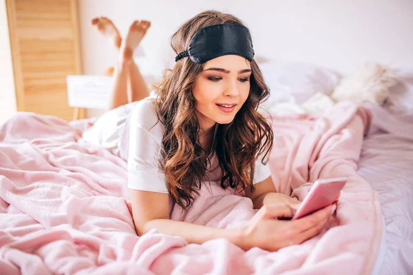 Mujer joven con el pelo oscuro acostado en la cama en el dormitorio solo. Cogiendo el teléfono en las manos y mirándolo. Usuario del dispositivo . —  Fotos de Stock