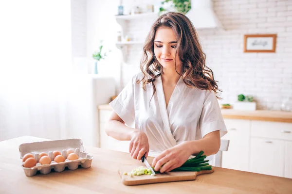 Eine junge Frau mit dunklen Haaren steht in der Küche und schneidet grüne Zwiebeln. Eier nebenbei. Morgendämmerung. Allein in der Küche. — Stockfoto