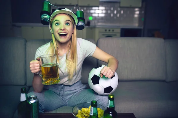Fiatal nő néz focimeccsen a tévében éjjel. Vidám vonzó szőke nő tart pohár sört és labdát. Palackok az asztalra. Ujjongott. Egyedül a szobán. — Stock Fotó