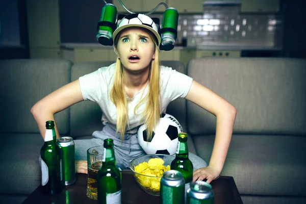 Young woman watch football game on tv at night. orried model look up forward. Alone in room. Bottles with beer on table. Ball between legs.