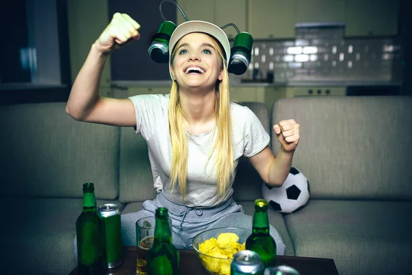 Ung kvinna titta på fotbollsmatch på TV på natten. Blond modell slå upp och leende. Streaming match på TV. Ölflaskor på bordet. Mörker i rummet. — Stockfoto