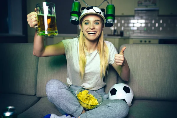 Young woman watch football game on tv at night. Positive attractive model look straight. Holding glass of beer and show big thumb up. Hat with beer cans on each side. Ball on sofa.