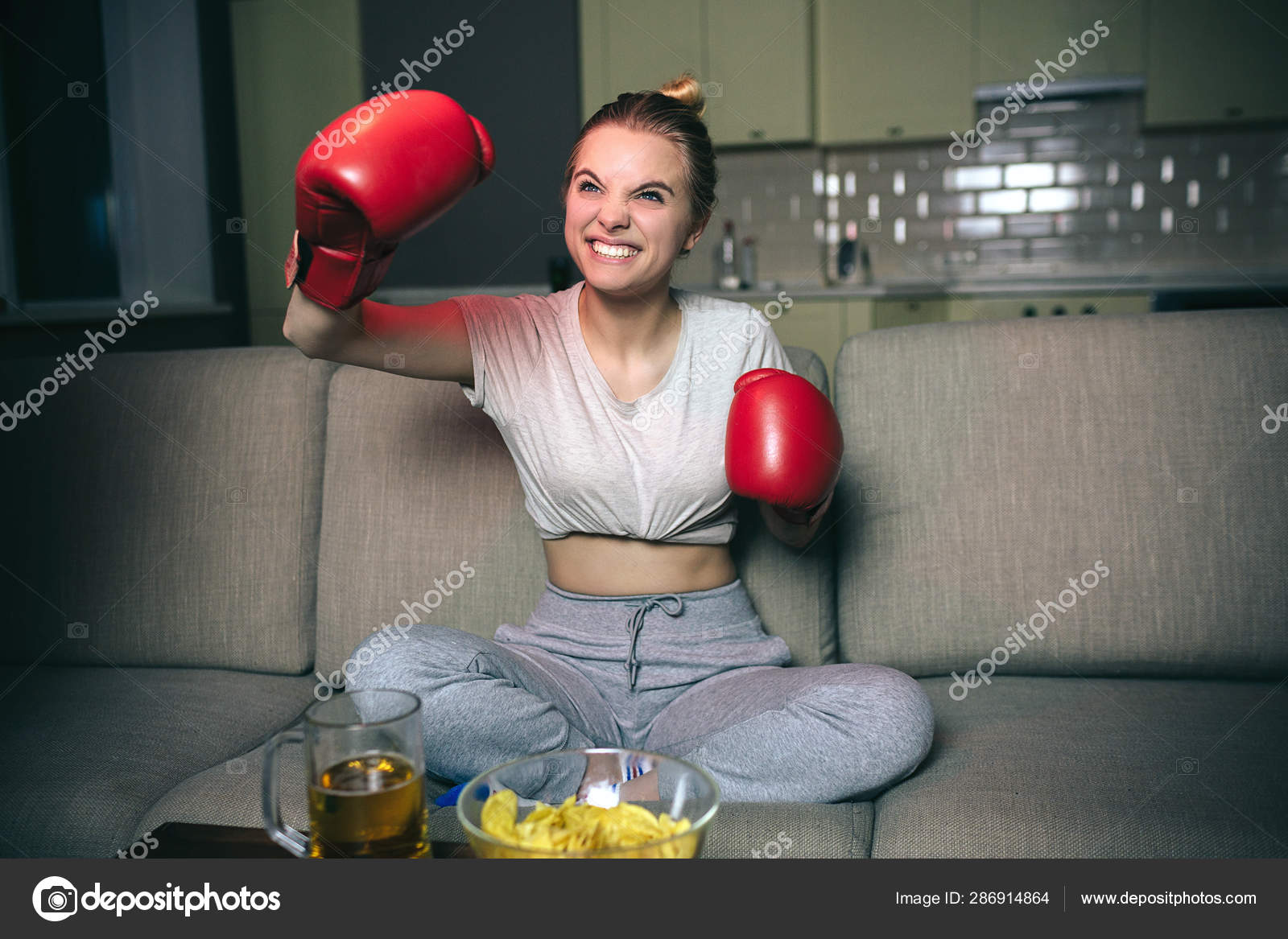 Young woman watch boxing on tv at night. Emotional blonde model cheering and wave with hands in red boxing gloves. Watching streaming show