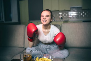 Genç kadın geceleri televizyonda boks izliyor. Duygusal kızgın model ileriye bakmak. Kızgın surat. Eller kırmızı spor eldivenler. Karanlık odada tek başına oturmak.