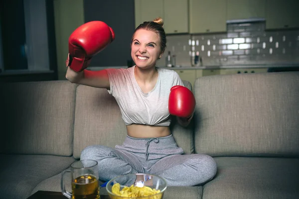 Mujer joven ver boxeo en la televisión por la noche. Emocional modelo rubia animando y saludando con las manos en guantes de boxeo rojos. Viendo el programa de streaming. Solo en la habitación . — Foto de Stock