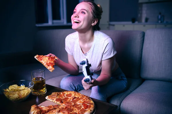 Young woman playing game at night. Happy positive model look up and smile. Holding gamepad and slice of pizza in hands. Junk food on table.