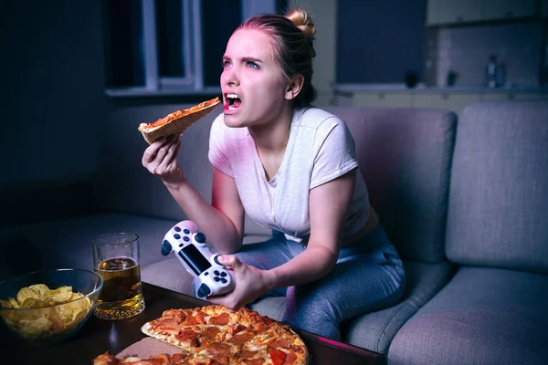 Young woman playing game at night. Emotional serious model eating slice of pizza. Game break. Holding gamepad in hand. Alone in dark room.