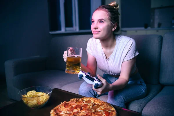 Jonge vrouw spelen spel 's nachts. Vrolijk aantrekkelijk model Drink bier en kijk uit. Junk food op tafel. Genieten van het spel. Gamepad in de hand. — Stockfoto
