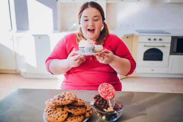Fat young woman in kitchen sitting and eating sweet food. Happy plus size model hold cakes in hands and smile. Body positive. Daylight in kitchen. — Stock Photo, Image
