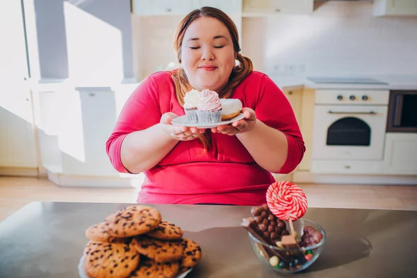 Mulher gorda na cozinha sentada e comendo comida doce. Além disso o modelo de tamanho mantém pequenos bolos em mãos e olha para eles. A mulher feliz gosta de comer. Corpo positivo . — Fotografia de Stock