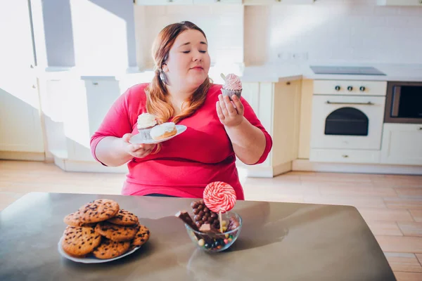 Mulher gorda na cozinha sentada e comendo comida doce. Além de tamanho modelo olhar para bolo na mão e sorrir. Prato com panquecas noutra. Corpo positivo. Bolachas na mesa. Luz do dia no quarto . — Fotografia de Stock