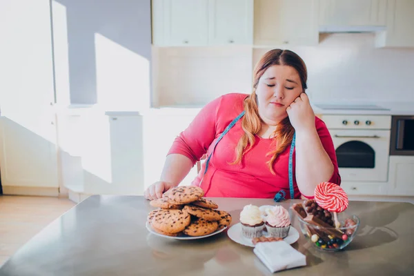 Mulher gorda na cozinha sentada e comendo comida doce. Aborrecido modelo plus size olhar para panquecas e doces na mesa. Luz do dia na cozinha. Corpo positivo. Fita métrica suave em torno do pescoço . — Fotografia de Stock