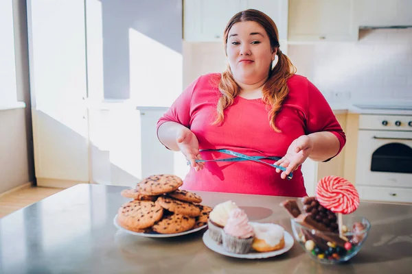 Mulher gorda na cozinha sentada e comendo comida doce. Fita métrica suave em torno da cintura e nas mãos. Além de olhar modelo de tamanho na câmera confusa. Bosy positivo. Doces e biscoitos à mesa. Luz do dia . — Fotografia de Stock