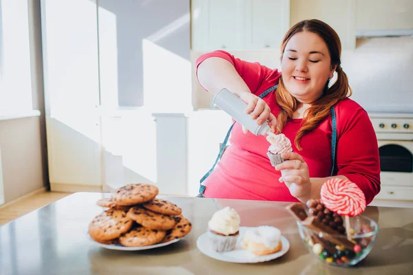 Χοντρή νεαρή γυναίκα στην κουζίνα κάθεται και τρώει γλυκό φαγητό. Ευτυχισμένο συν μέγεθος μοντέλο βάλει λίγη κρέμα στην τηγανίτα. Το σώμα είναι θετικό. Μπλε μαλακό μεζούρα. Γλυκά και μπισκότα στο τραπέζι. Θερινή ημέρα — Φωτογραφία Αρχείου