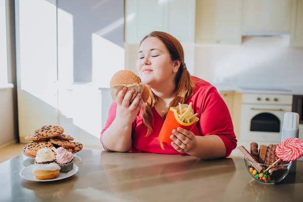 Mulher gorda na cozinha sentada e comendo junk food. Estilo de vida pouco saudável. Segure hambúrguer e batatas fritas nas mãos. Corpo positivo. Doces na mesa. Luz do dia na cozinha . — Fotografia de Stock