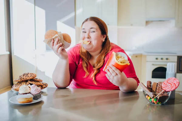 Mulher gorda na cozinha sentada e comendo junk food. Batatas fritas na boca. Olhando para a câmera e sorrir. Corpo positivo. Hambúrgueres nas mãos. Estilo de vida pouco saudável . — Fotografia de Stock