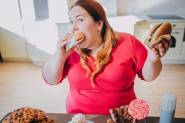 Fat young woman in kitchen sitting and eating junk food. Unhealthy lifestyle. Biting piece of burger and look on camera. Body positive. Sweets on table. Daylight in kitchen.