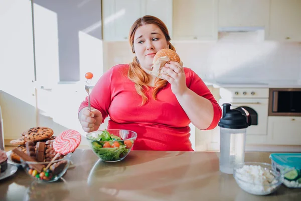 Χοντρή νεαρή γυναίκα στην κουζίνα κάθεται και τρώει πρόχειρο φαγητό. Συν μέγεθος μοντέλο έχει ανθυγιεινό φαγητό. Το μπολ με τη σαλάτα στο τραπέζι. Το σώμα είναι θετικό. Ένα κομμάτι μπέργκερ. Δίαιτα. — Φωτογραφία Αρχείου