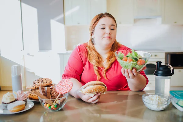 Mulher gorda na cozinha sentada e comendo comida. Tigela com salada na mão. Além disso, o modelo de tamanho olhar para ele. Corpo positivo. Jovem confuso na cozinha . — Fotografia de Stock