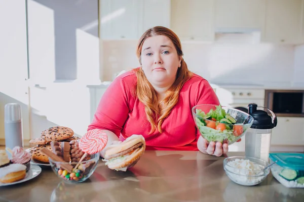 Mulher gorda na cozinha sentada e comendo comida. Confuso infeliz plus size modelo segurar hambúrguer e salada tigela nas mãos e olhar para a câmera. Sozinho na cozinha . — Fotografia de Stock
