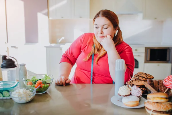 Mulher gorda na cozinha sentada e comendo comida. Ela olha para uma refeição saudável do lado esquerdo. Fita métrica suave em torno do pescoço. Comida de plástico doce do lado direito. Luz do dia na cozinha . — Fotografia de Stock