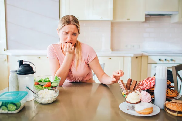 Молодая несчастная стройная модель трогает печенье на кухне. Она смотрит на фаст-фуд со стола. Здоровое питание с левой стороны. Один и грустно . — стоковое фото