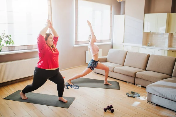 Youbng modelo de talla grande y mujer bien construida haciendo ejercicio juntos. Estirarse y pararse en postura de yoga. Meditación Asana. Entrenamiento para dos en sala de estar . — Foto de Stock