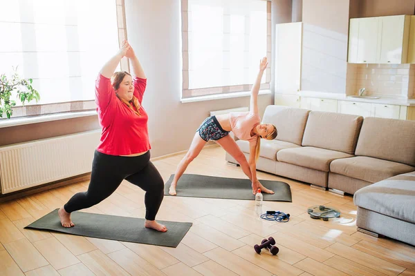 Jóvenes delgados y con sobrepeso modelos exersicing juntos en la sala de estar. De pie en asana o posturas de yoga. Entrenamiento para dos. Paraguas en el suelo . — Foto de Stock