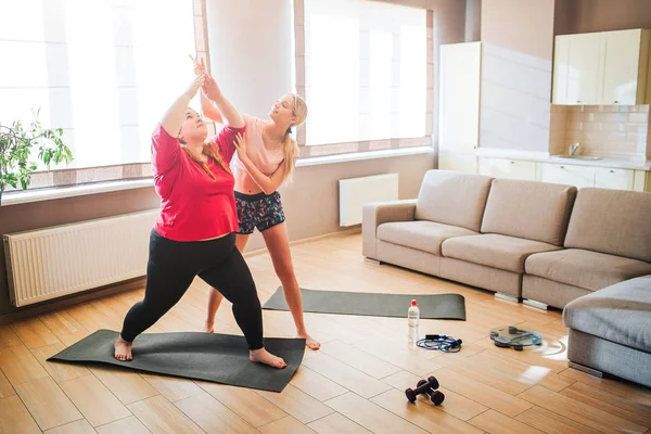 Joven mujer delgada bien construida ayuda más modelo de tamaño para estirar y hacer ejercicio en la sala de estar. Mirando hacia arriba. Sufrir de entrenamiento duro. Luz del día . — Foto de Stock