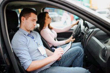 Male auto instructor takes exam in young woman. Busy, serious and concentrated man writing test results on paper. Confident female driver look forward on road. Passing driving exam for getting lisence