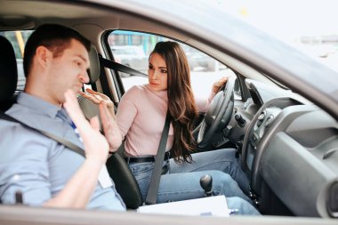 Male auto instructor takes exam in young woman. Female student look at teacher with confusion and misunderstanding. Guy show hands and look down. Daylight.