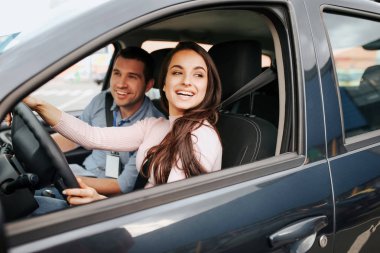 Male auto instructor takes exam in young woman. Cheerful attractive female driver posing at drivers place. Look from car. Hold hands on sheering wheel.
