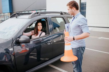 Male auto instructor takes exam in young woman. Stand outside of car with orange sign in hands. Look at woman in car. Female studenthold hands on steering wheel adnd look at instructor.