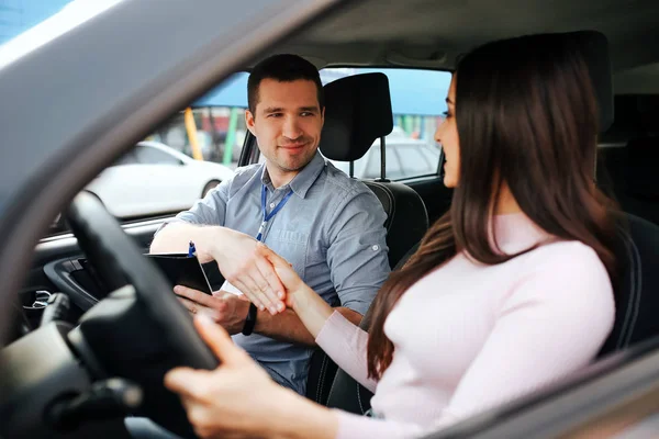 Male auto Instructor neemt examen in jonge vrouw. Handen schudden. Zittend in de auto. Brunette houden hand op stuurwiel. — Stockfoto